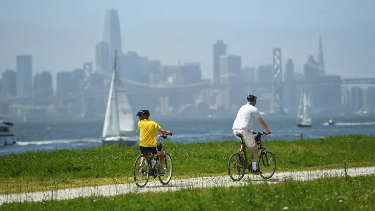 Two people riding bikes on a dirt path in Middle Harbor Shoreline Park, with a view of sailboats, the San Francisco-Oakland Bay Bridge and the San Francisco downtown skyline in the background.