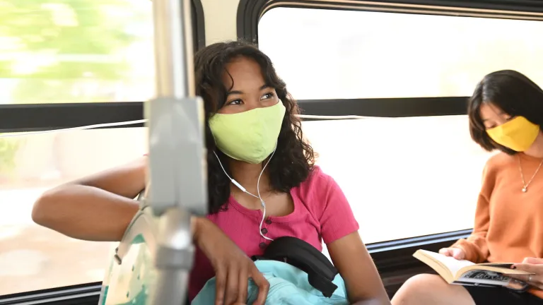 A young woman wearing a mask and earphones looks out the window while riding a bus.
