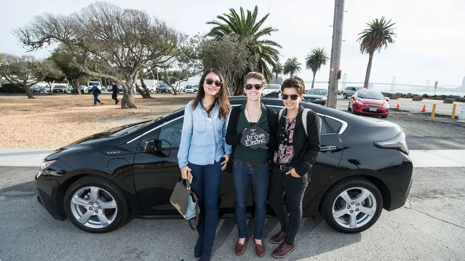 three women posing in front of electric car